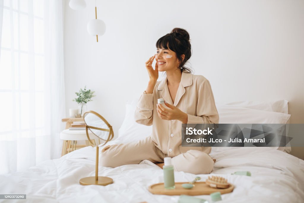 Happy woman doing routine skin care at home with beauty products. Woman sitting on bed at home and applying face cream.