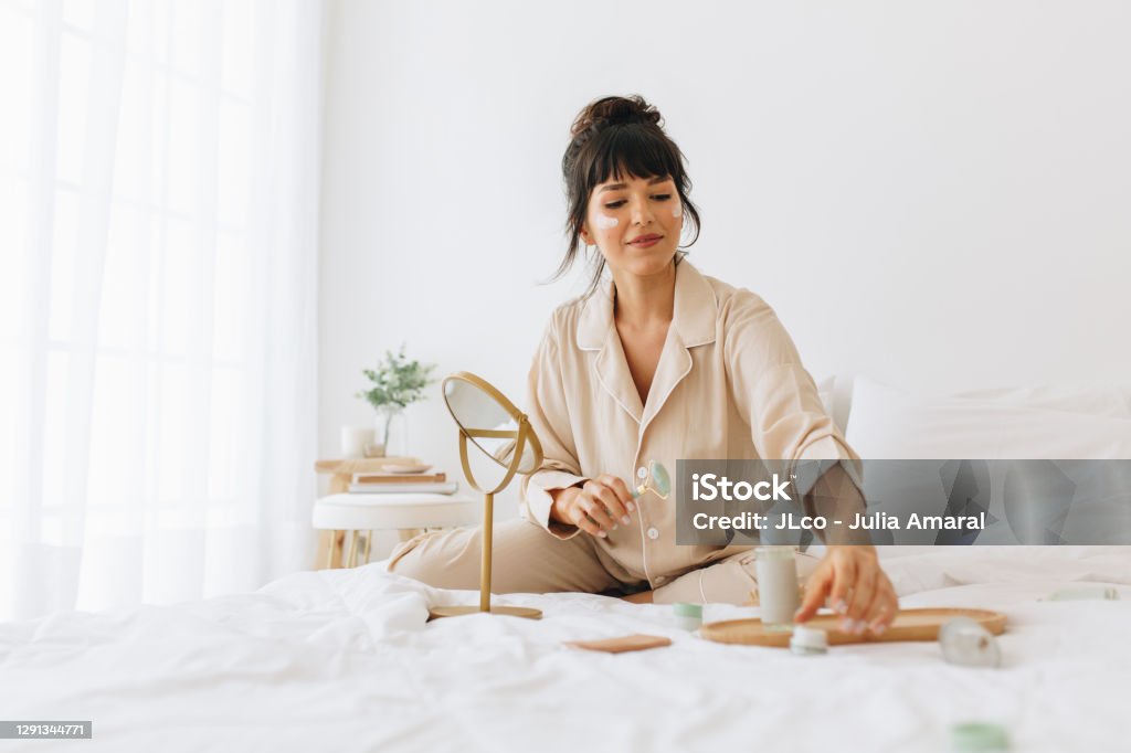 Young woman sitting on bed at home and applying cosmetics. Woman using skin care products at home.